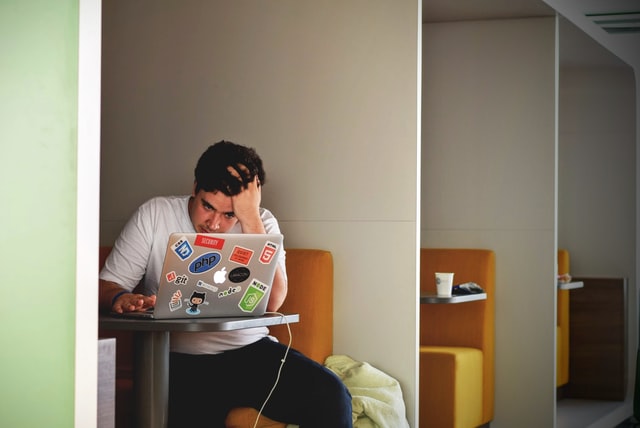 Homem de calça preta e camiseta branca, sentado à mesa de trabalho no laptop com adesivos.